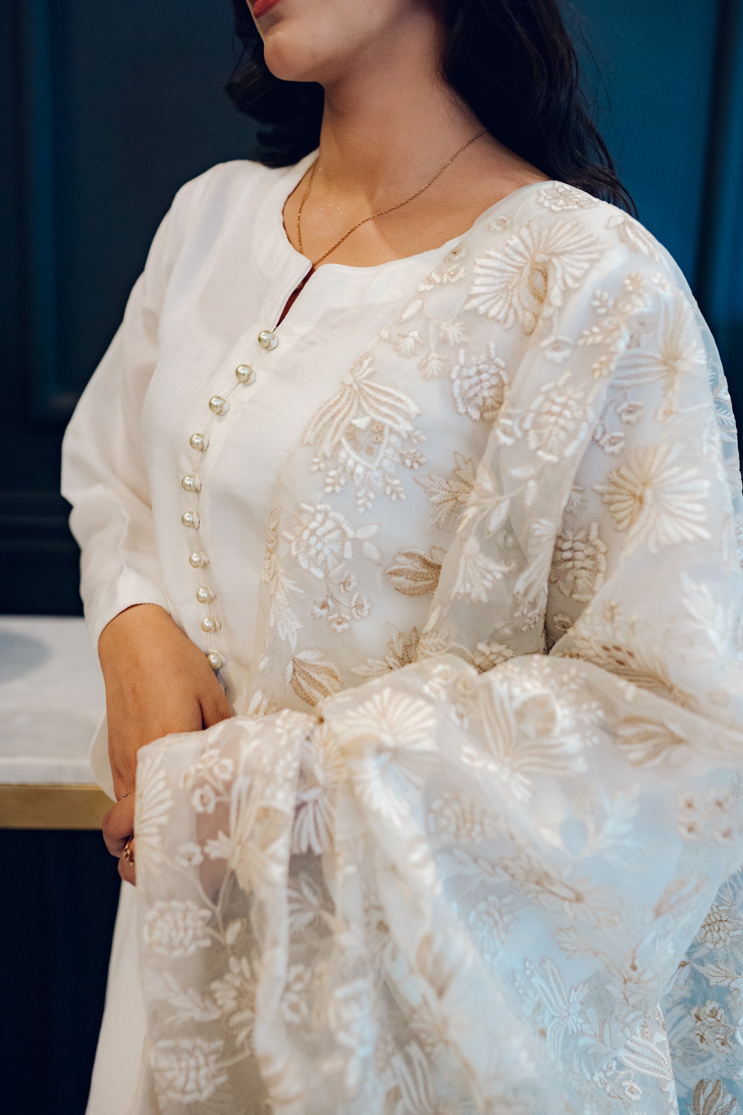 Ivory dress featuring delicate pearl buttons, paired with a shimmering tila and intricate threadwork dupatta, showcasing timeless elegance and artisanal craftsmanship.