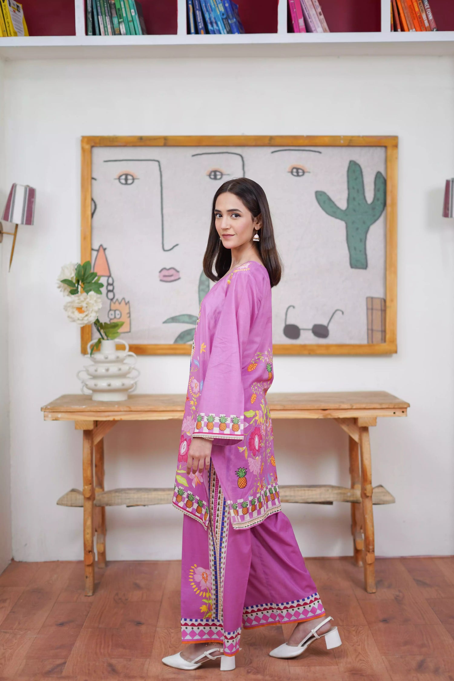Woman in a pink traditional outfit standing in a room with a wooden table and framed artwork.