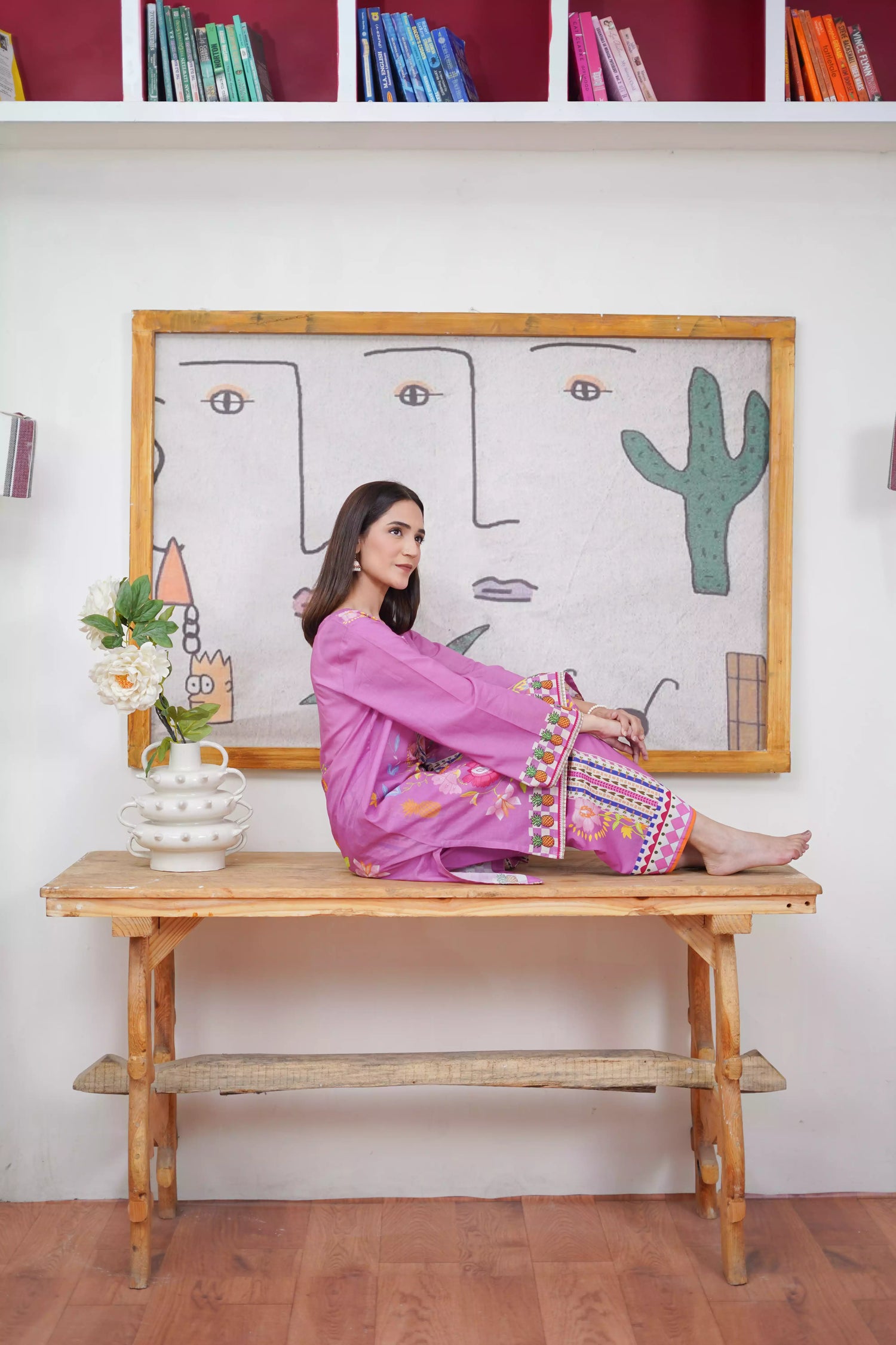 Woman in a pink dress sitting on a wooden table with a colorful abstract painting in the background.